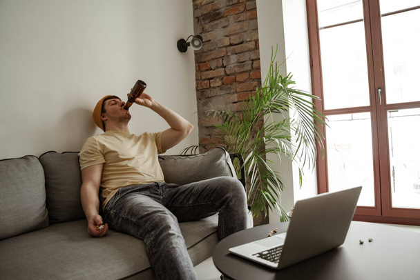 Joven turista en sombrero amarillo bebiendo cerveza con portátil en la habitación de hotel, hombre de vacaciones - Foto, Imagen