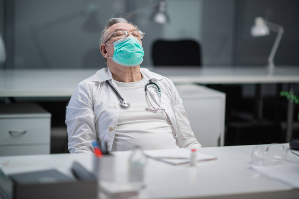 医療マスクの高齢者の経験豊富な医者が彼のオフィスに戻って、彼は流行中に仕事の一日後に非常に疲れていた - 写真・画像