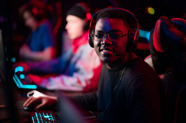 Neon light in the arena at the online shooter tournament. A young black cyber athlete looks at the camera. Blurred background. - Photo, Image