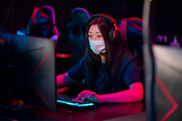 Um jovem asiático menina vestindo uma máscara joga jogos de vídeo online à noite em um clube de computador - Foto, Imagem