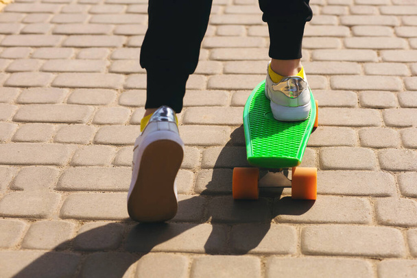 Een kind rijdt op een groen skateboard of penny board in het park. Het meisje is gekleed in heldere kleren en houdt van skateboarden. De gezondheidsvoordelen van buitensporten. Skateboard en benen bekijken - Foto, afbeelding