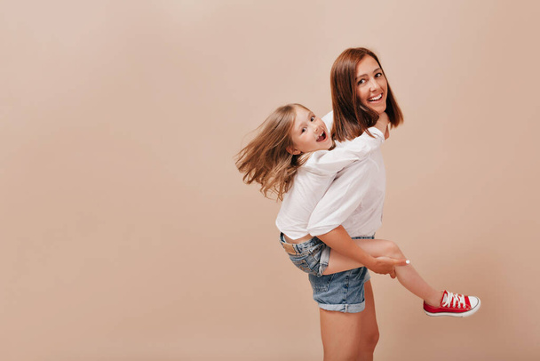 Lovely little girl in white shirt and jeans smiling with happy smile and hugging her mother in stylish clothes on beige backdrop.. High quality photo - Fotografie, Obrázek