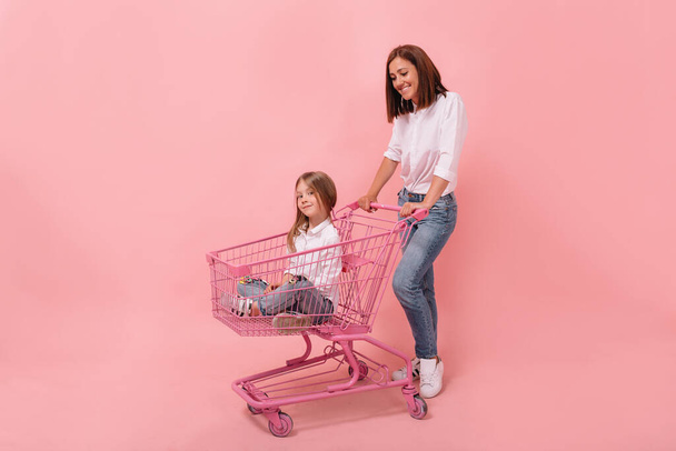 Giovane donna in camicia bianca che gioca con la sua figlioletta su sfondo rosa. Ragazzina seduta nel cestino rosa con sua madre. Foto di alta qualità - Foto, immagini
