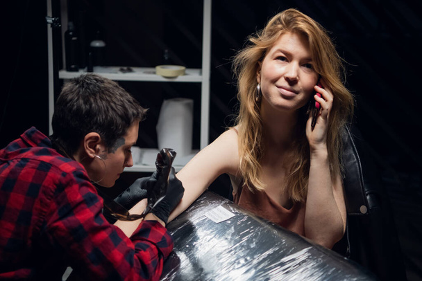 Um tatuador experiente com um rosto tatuado enche uma tatuagem para seu cliente uma garota com cabelo vermelho enquanto ela está falando em um smartphone. - Foto, Imagem