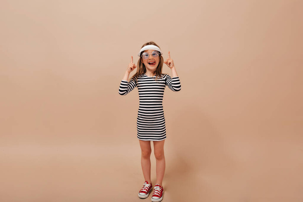 Φωτογραφία του γοητευτικού κοριτσιού με καπέλο και κόκκινα sneakers που ποζάρει με χαμόγελο και κρατάει ψηλά τα δάχτυλα. Υψηλής ποιότητας φωτογραφία - Φωτογραφία, εικόνα
