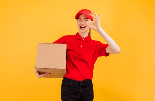 Παράδοση σε κόκκινη στολή κρατώντας ένα δέμα χαρτοκιβωτίων, Γυναίκα υπάλληλος που εργάζεται ως κούριερ, σε κίτρινο φόντο, Υπηρεσία έννοια - Φωτογραφία, εικόνα