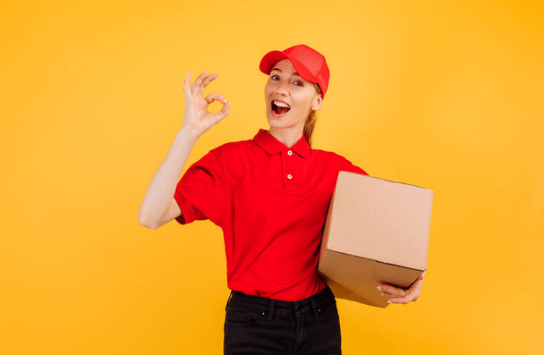 Delivery in red uniform holding a carton box parcel, Female employee working as a courier, on a yellow background, Service concept - Φωτογραφία, εικόνα