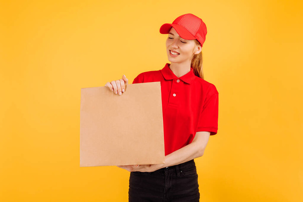 Delivery service employee in a red cap and t-shirt, holding a paper bag with food on an isolated yellow background - Valokuva, kuva