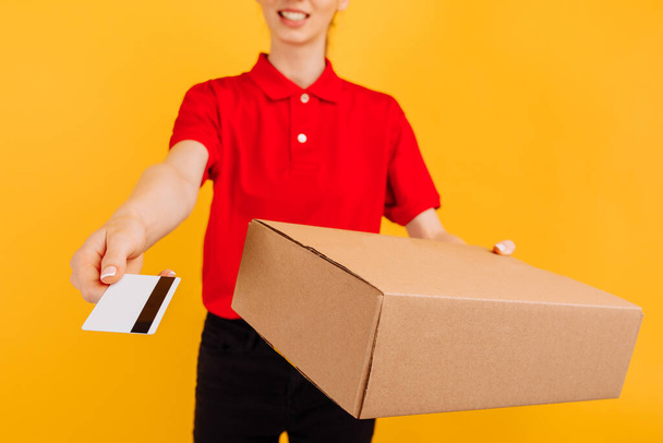 Happy young woman courier, in red uniform, delivery officer holding cardboard box and credit card on isolated yellow background - Фото, зображення