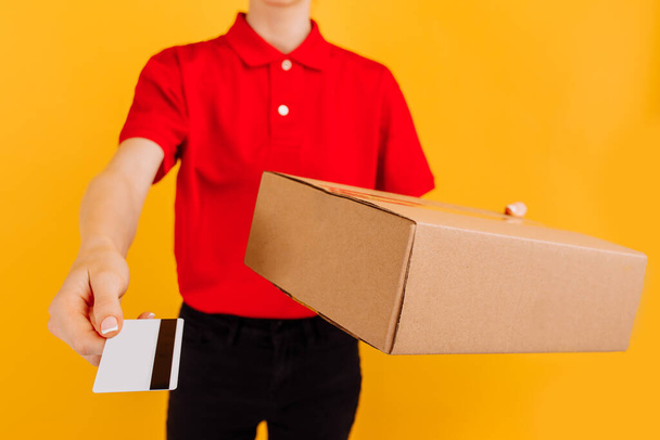 Щаслива молода жінка кур'єр, в червоній формі, службовець доставки тримає картонну коробку і кредитну картку на ізольованому жовтому фоні
 - Фото, зображення