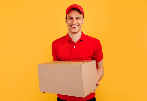 Image of happy young courier in red hat standing with mailbox isolated on yellow background - Fotoğraf, Görsel