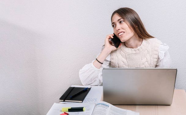 Νεαρή γυναίκα χαμογελά ενώ εργάζεται στο σπίτι χρησιμοποιώντας τον υπολογιστή και μιλώντας στο κινητό. Έννοια επιχειρηματία. Έννοια της ελευθερίας. Εργασία στο σπίτι. - Φωτογραφία, εικόνα