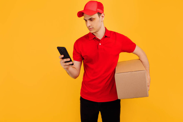 Delivery service employee in a red cap and t-shirt, uses a mobile phone and holds a parcel on a yellow background - Photo, image