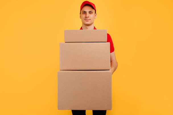 Toimitus työntekijä, mies kuriiri punainen t-paita ja lippis, jossa joukko pahvilaatikoita eristetty keltainen tausta - Valokuva, kuva