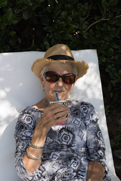 alacsony ősz hajú, felnőtt nő napszemüveggel és sapkával. Az öreg hölgy iszik, és egy nyugágyon ül a háza kertjében. Az ősi fekete-fehér pólót visel, és a kamerába néz. a nap süt, így van - Fotó, kép