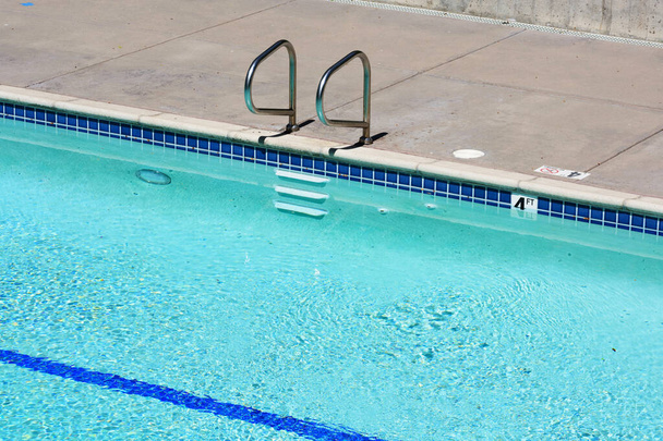 Zvýšený výhled na venkovní bazén s kovovými madly se schody, značkovač hloubky na okraji bazénu, podvodní svítidlo a modrá čára na dně bazénu - Fotografie, Obrázek