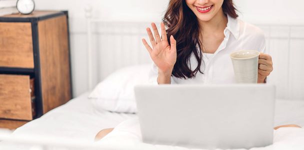 Young smiling asian woman relaxing using laptop computer and drinking coffee in bedroom at home.Young creative girl working and video conference online meeting with colleagues.work from home concept - Фото, изображение
