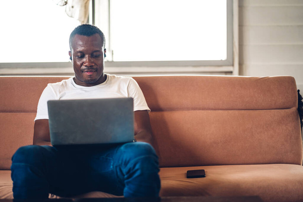 Молодой чернокожий африканский мужчина, расслабляющийся с помощью ноутбука, работающего на компьютере, и видеоконференции в доме. - Фото, изображение