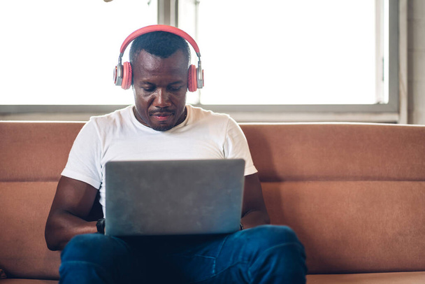 Genç siyahi Afrikalı adam dizüstü bilgisayarla evde çalışıyor ve video konferansı yapıyor. Genç yaratıcı Afrikalı adam kulaklıkla konuşuyor. Ev konseptinden çalışma. - Fotoğraf, Görsel