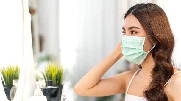 Young asian woman clean fresh healthy white skin looking at mirror in quarantine for coronavirus wearing surgical mask face protection with social distancing at home.covid19 and new normal concept - Photo, Image