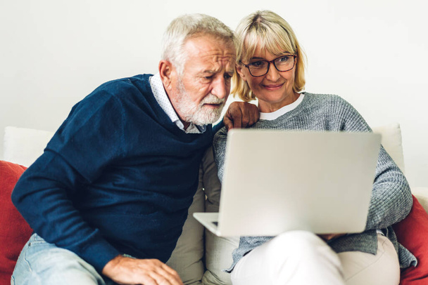 一緒にラップトップコンピュータを使用して楽しい時間を過ごしているシニア夫婦。幸せな高齢者の夫と妻ソーシャルメディアをチェックし、自宅でソファに座っている間にニュースやショッピングをオンラインで読む - 写真・画像