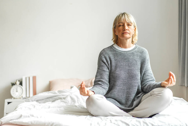 Portrét starší ženy sedící a dělat jógu. Atraktivní starší žena fitness cvičení cvičení na posteli doma - Fotografie, Obrázek