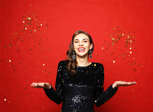 Holiday and party concept: beautiful young woman wearing evening dress standing under confetti rain over red background - Photo, image