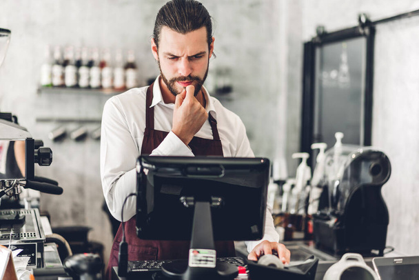 Portret van een knappe barista man met baard, kleine ondernemer, werkend met laptop achter de bar in een café - Foto, afbeelding