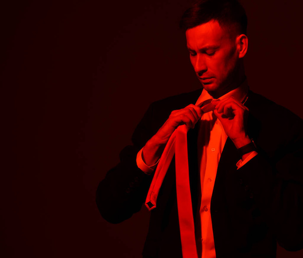Portrait of young focused businessman in official wear, suit tying his tie looking at it. Red light filter, gel - Foto, Imagem