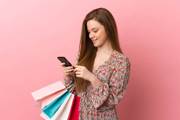 Ragazza adolescente su sfondo rosa isolato tenendo borse della spesa e scrivendo un messaggio con il suo telefono cellulare ad un amico - Foto, immagini