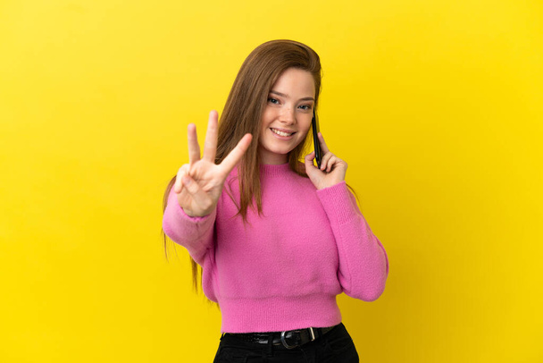 Adolescente chica usando el teléfono móvil sobre aislado amarillo fondo feliz y contar tres con los dedos - Foto, Imagen
