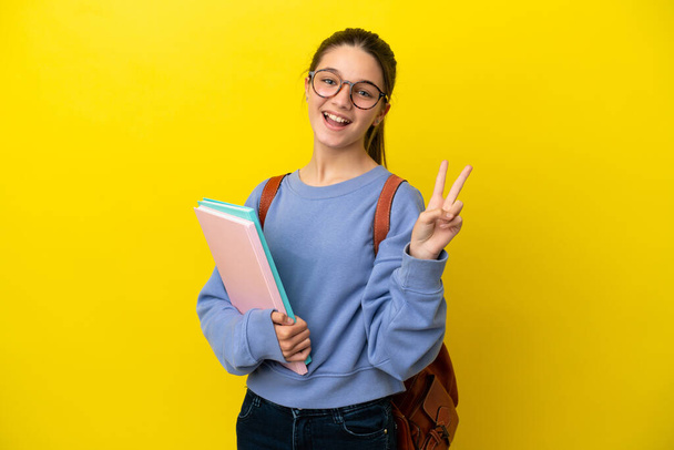 Студентка девочка на изолированном желтом фоне улыбается и показывает знак победы - Фото, изображение