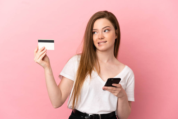 Adolescente chica sobre aislado rosa fondo compra con el móvil con una tarjeta de crédito, mientras que pensando - Foto, Imagen