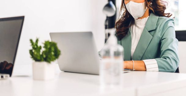 オフィスの机の上に座っている間、社会的距離と保護マスクを身に着けているコロナウイルスの隔離のためのラップトップコンピュータの作業と計画会議を使用して若いアジアのビジネス女性 - 写真・画像