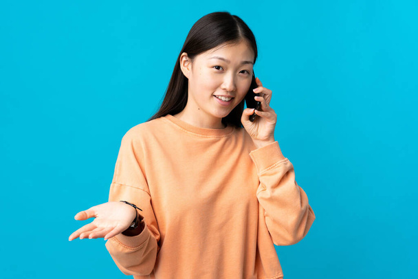 Νεαρή Κινέζα πάνω από απομονωμένο μπλε φόντο κρατώντας μια συνομιλία με το κινητό τηλέφωνο με κάποιον - Φωτογραφία, εικόνα