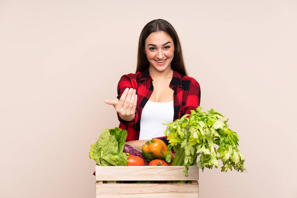 Farmer with freshly picked vegetables in a box isolated on beige background inviting to come - Photo, Image