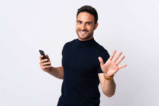 Jeune homme caucasien utilisant téléphone portable isolé sur fond blanc saluant avec la main avec expression heureuse - Photo, image