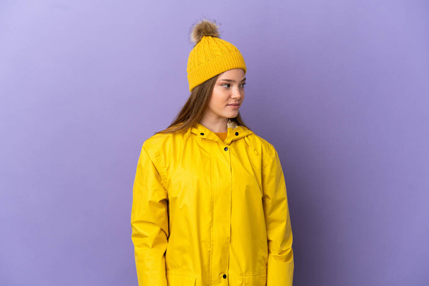 Tini lány visel esőálló kabát felett elszigetelt lila háttér néz oldalra - Fotó, kép