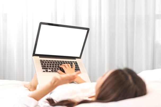 自宅のベッドの上に白いモックアップの空白の画面を持つラップトップコンピュータの技術を使用してリラックスした女性。 - 写真・画像