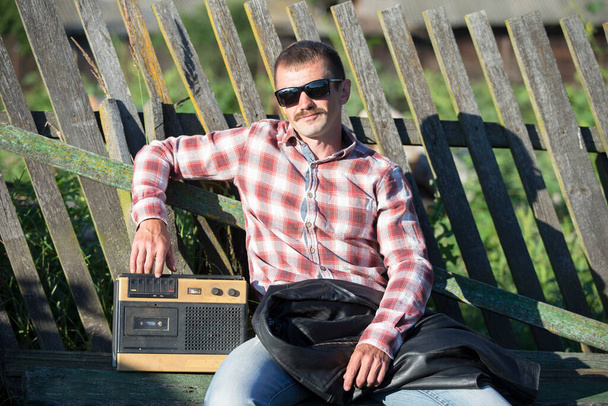 Un hombre gracioso con una grabadora de casetes se sienta junto a una valla de madera. - Foto, Imagen