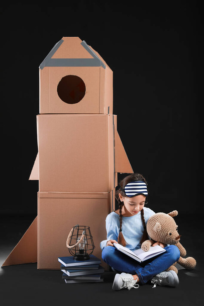 Cute little girl with cardboard rocket, books and toy on dark background - Photo, Image