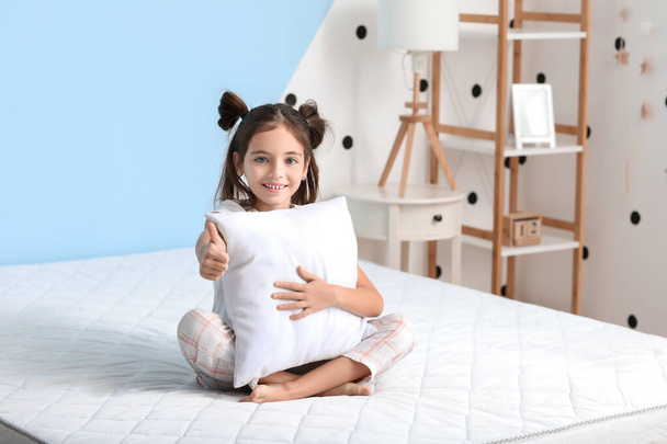 Morgen eines kleinen Mädchens, das den Daumen nach oben zeigt, während es mit einer bequemen Matratze auf dem Bett sitzt - Foto, Bild