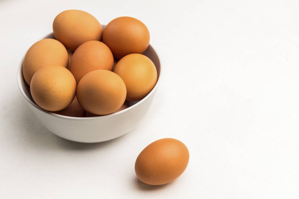 Brown eggs in white ceramic bowl. One egg on table. Copy space. White background. Top view - Φωτογραφία, εικόνα