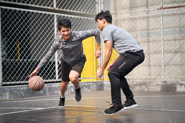 два молоді азіатські дорослі чоловіки грають один на один баскетбол на відкритому майданчику
 - Фото, зображення