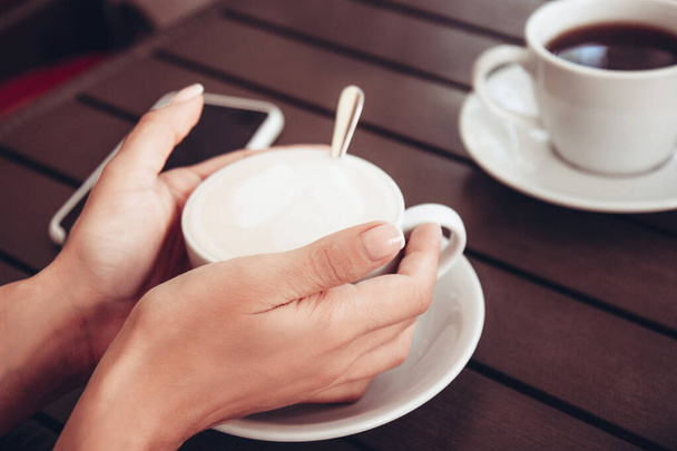 Filiżanka cappuccino ze sztuką latte w pięknych kobiecych dłoniach i filiżanka czarnej aromatycznej kawy na drewnianym stole. Biała ceramiczna filiżanka kawy. Rustykalna kawa na świeżym powietrzu - Zdjęcie, obraz