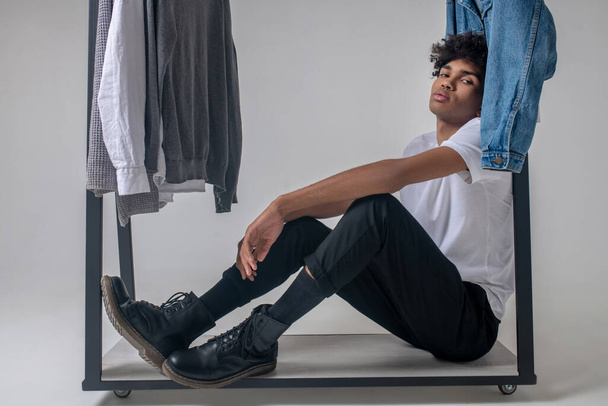 Curly-haired young guy sitting near the hangers with clothes and looking tired - Foto, imagen