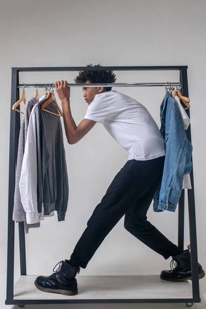 African american young guy holding hangers with clothes and leaning forward - Foto, afbeelding