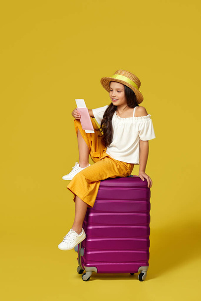 child girl in hat with passport and ticket sitting on suitcase - Фото, изображение