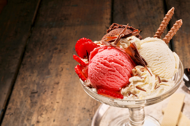 Gourmet-Eis aus Schokolade, Erdbeere und Vanille, garniert mit frischen Früchten und garniert mit Schlagsahne und Spezialitäten-Keksen vor rustikalem Holzhintergrund - Foto, Bild