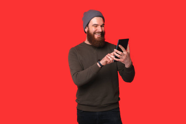 Cheerful young bearded hipster man using tablet over red background. - Zdjęcie, obraz